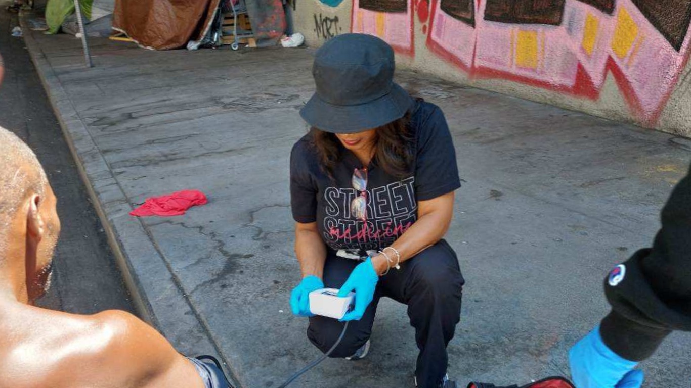 Health Matters Clinic clinician kneeling on the pavement to take a patient reading beside an encampment